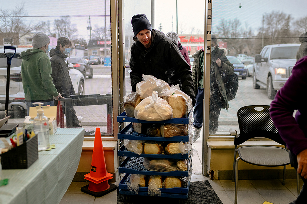 Chris, who is wearing a black jacket and toque, pushes a multi-tiered cart containing bagged loaves of bread into a food bank.