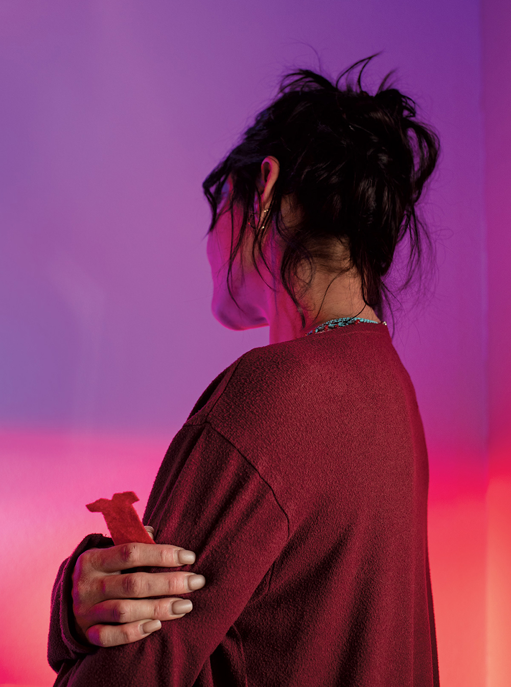 Angled back view of Chelsea in a red cardigan, her right hand clasping a small orange dress cut-out and holding her left arm