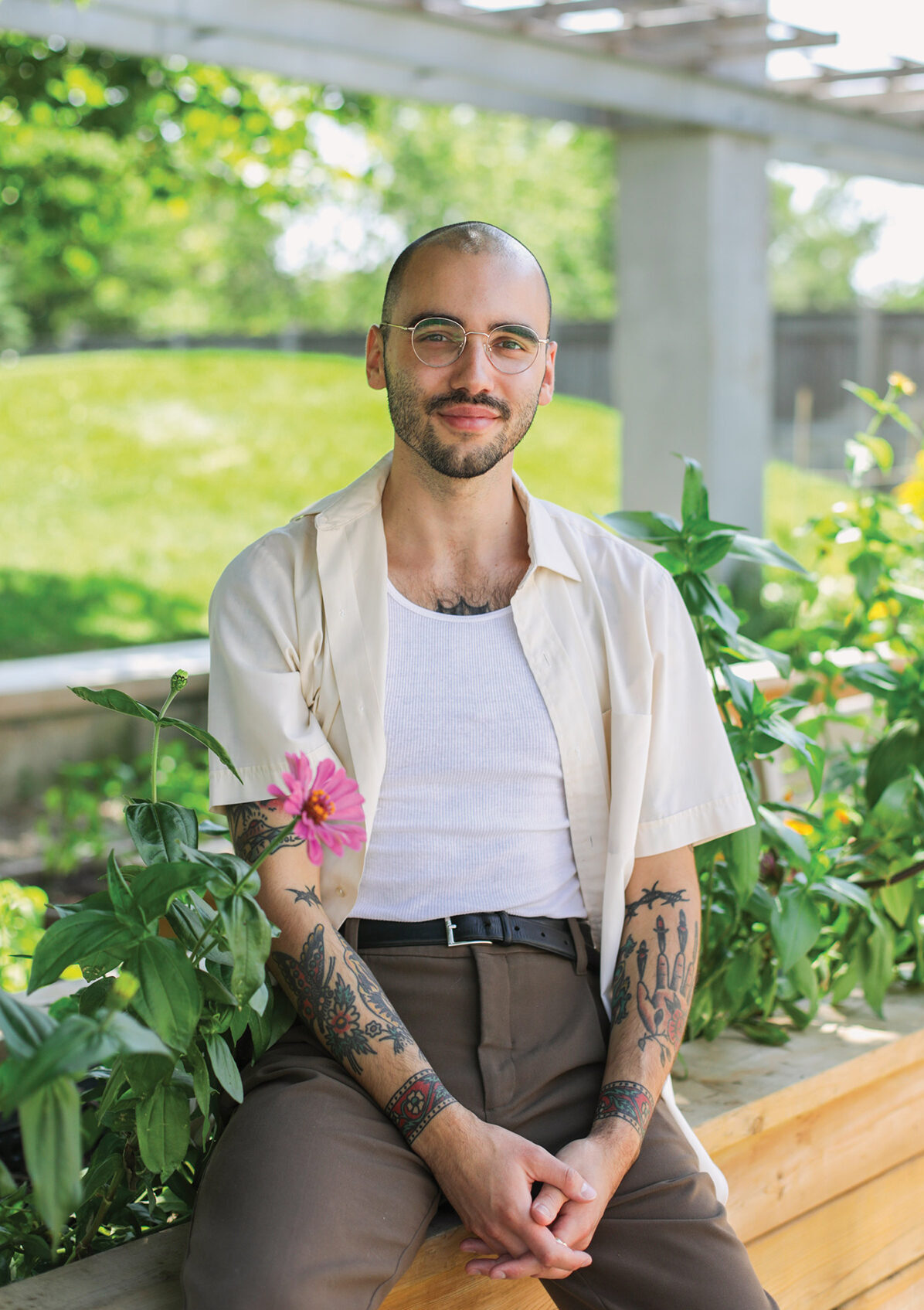 Outdoor photo of Cody Caetano in a white T-shirt under an off-white short-sleeved button-up shirt, revealing tattoos along his forearms. He is seated on a wooden bench with a flower bed.