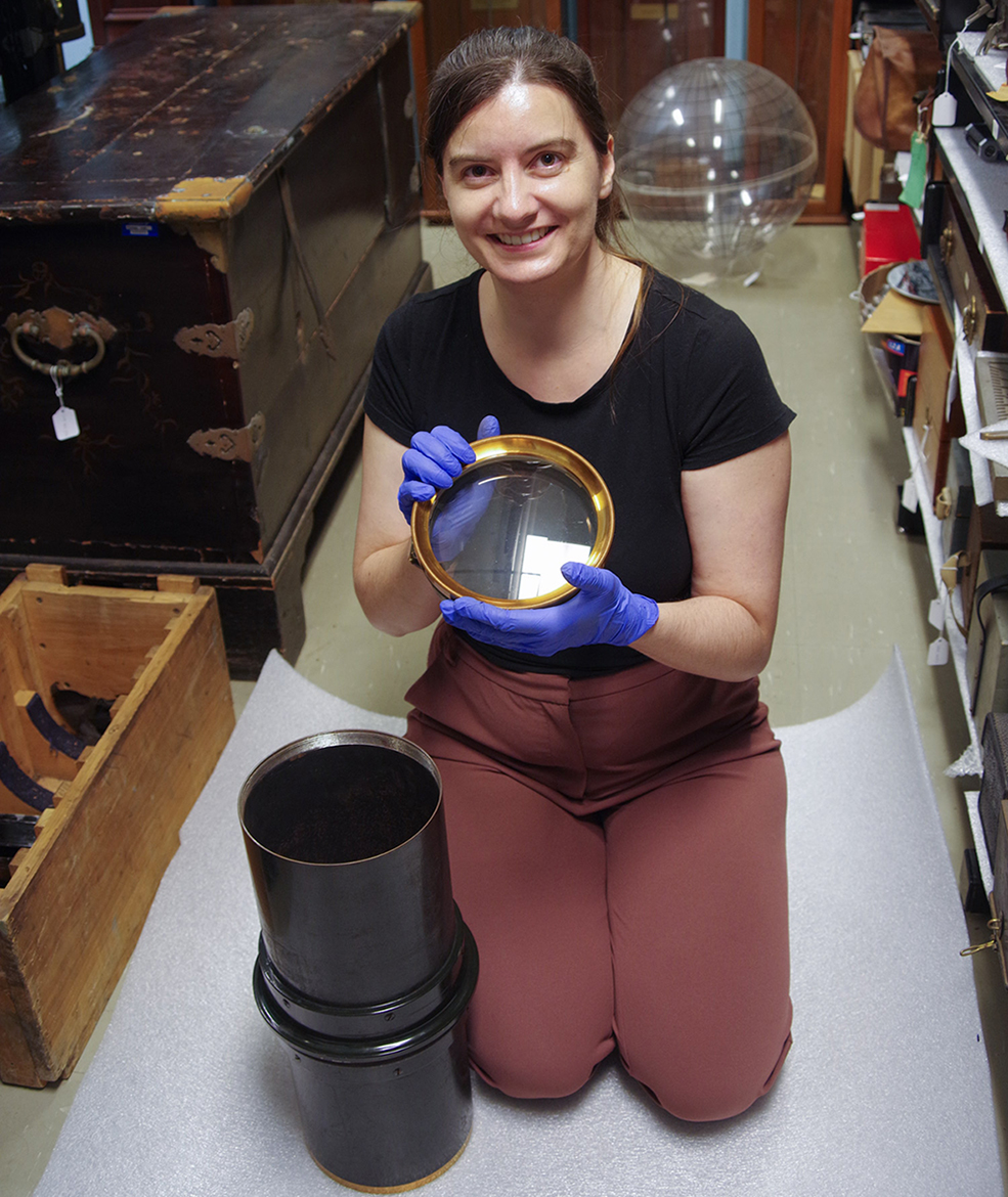 A woman, wearing a black T-shirt, mauve pants and blue disposable gloves, is kneeling on the floor of a storage room, holding a large round brass-framed lens. A black metal tube is placed upright on the floor beside her knee.