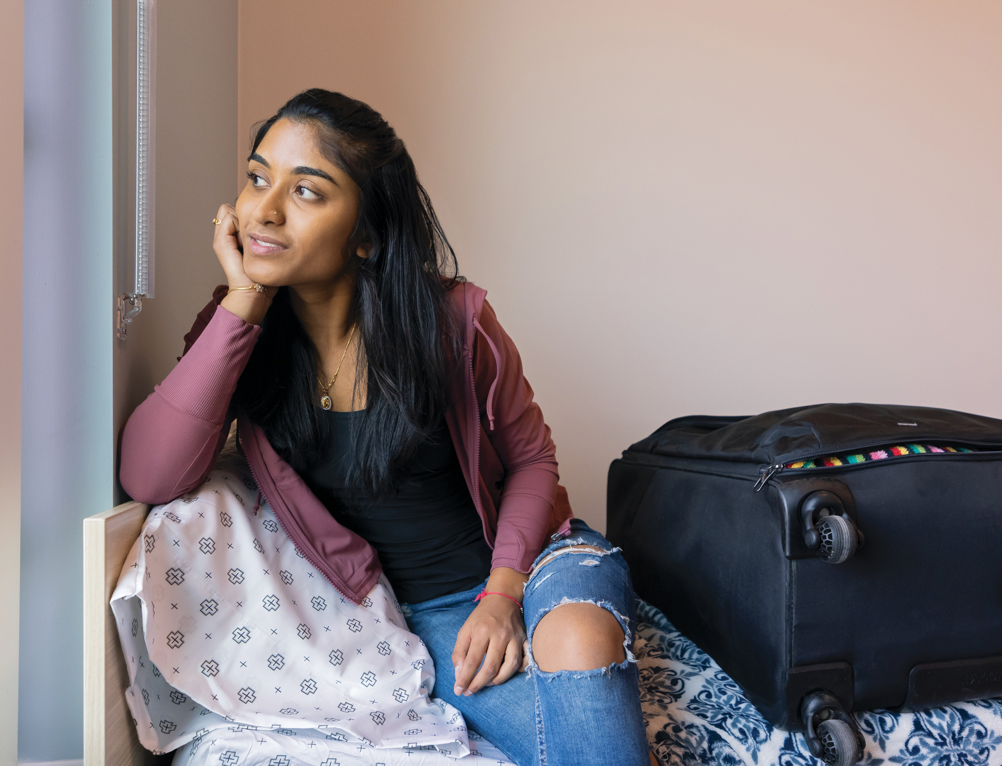 First-year student Reva Birla, in a black shirt, mauve hoodie and ripped blue jeans, is sitting on the side of her bed next to a large, black suitcase and looking out the window of her dorm room.