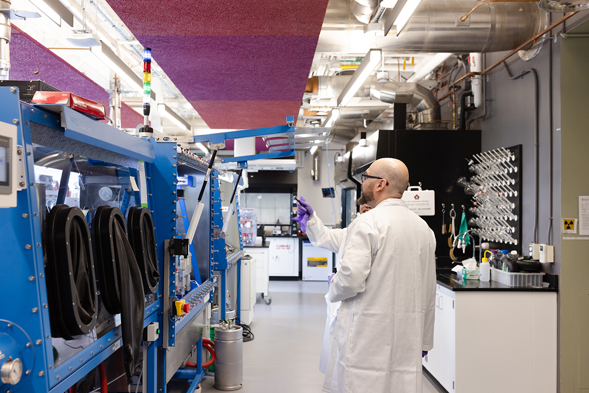 A bald man in a white lab coat stands mid frame. He's talking to someone next to him, but we can only see his purple lab gloves. To their left is a large blue container and a glass covering. There are three large oval shaped openings with black edges that allow researchers to get inside the container, presumably to do experiments