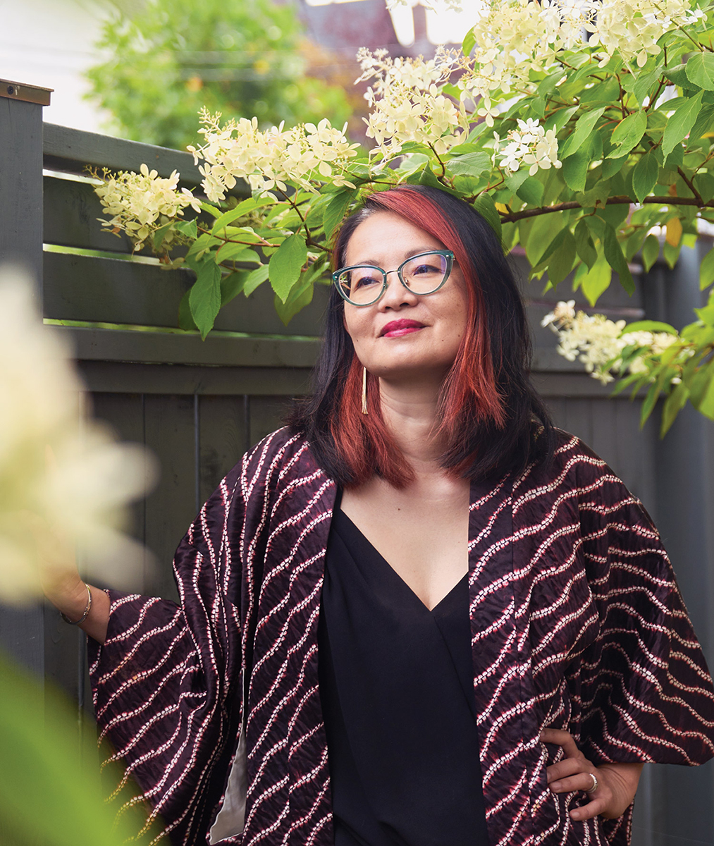 Professor Thy Phu, with thick blue-framed glasses and the front strands of her black hair dyed in red, stands with one hand on her hips and the other resting against a tall grey wooden fence. Behind and above her is a tree branch laden with leaves and white flowers.