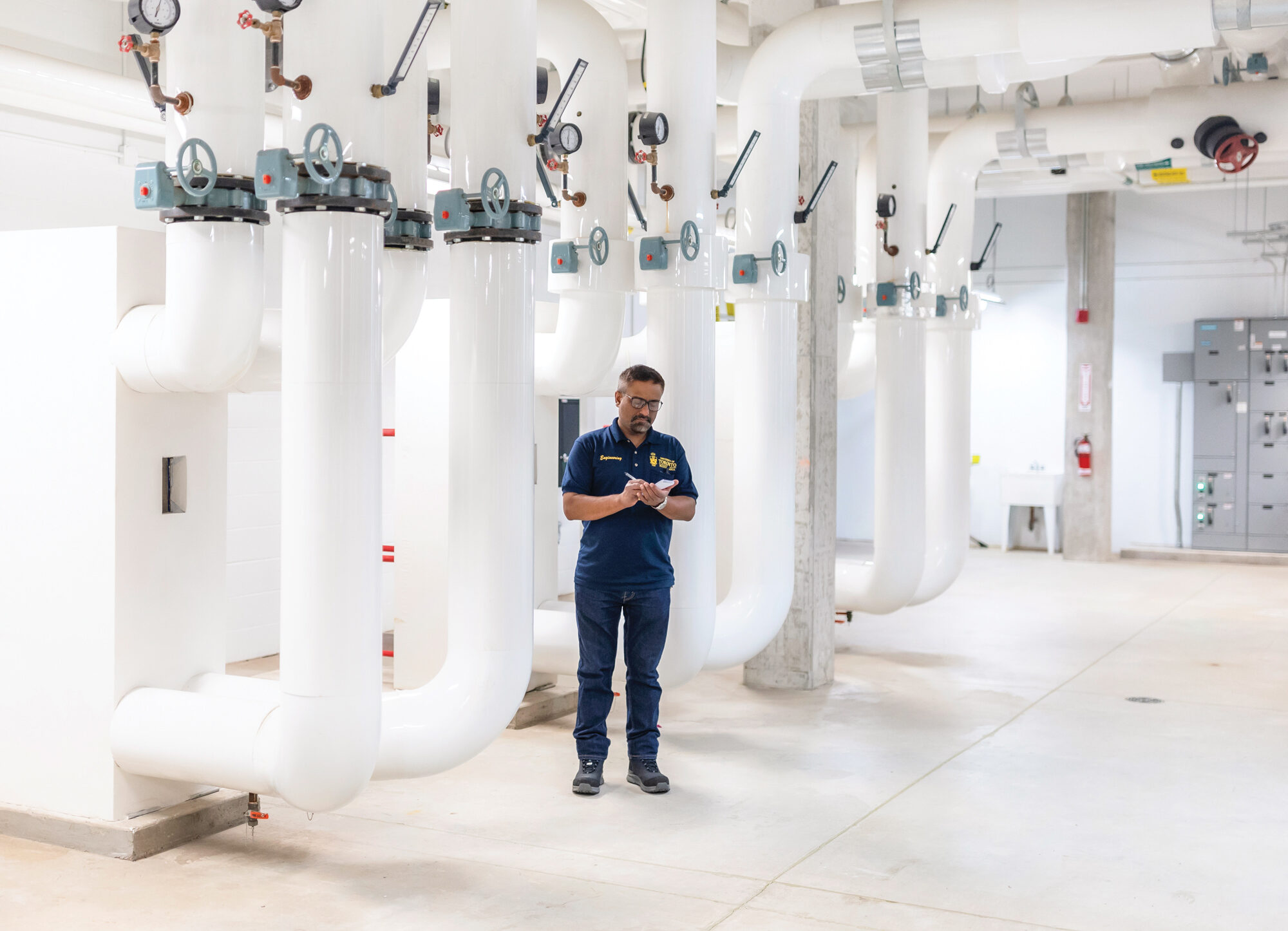 Building engineer Gurtaj Bajwa is writing on a small notepad in a room with large white pipes running parallel to the walls and ceilings, housing the geothermal system at U of T Scarborough's Instruction Centre