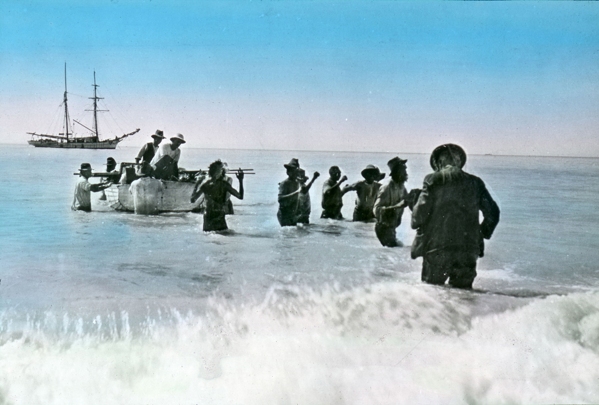 A group scientists are disembarking from a whaleboat, thigh deep in the surf. A few Nyangumarta people are assisting the scientists with carrying equipment from the boat. In the distance, a schooner sits at anchor