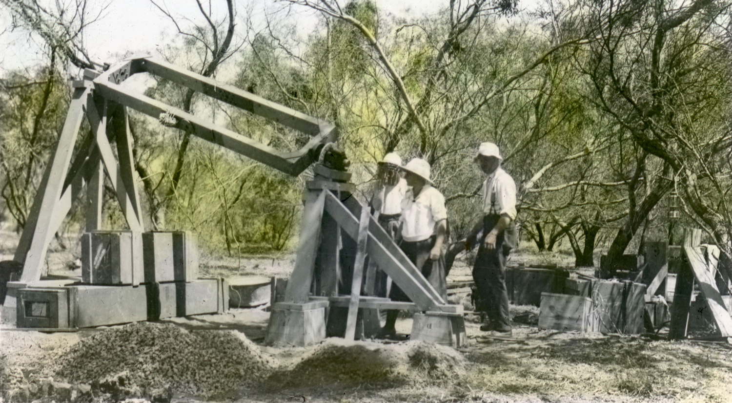 A wooden structure made of beams and rotational elements are set on the ground in a wooded area with short, sparse trees and an open sky. Three scientists wearing white hats are standing next to the structure.