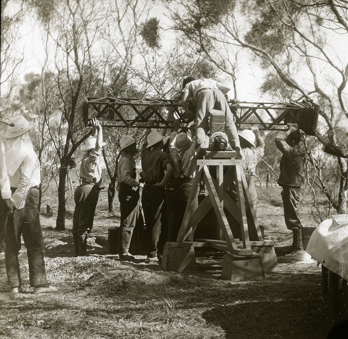 A sepia coloured photo of a group of scientists in white hats, holding over their heads a long metal tube-like structure with the photographic plate holder on one end. One scientist is helping with positioning the metal tube, while standing on top of a tall wooden tripod-like structure, where part of the Einstein camera is placed.