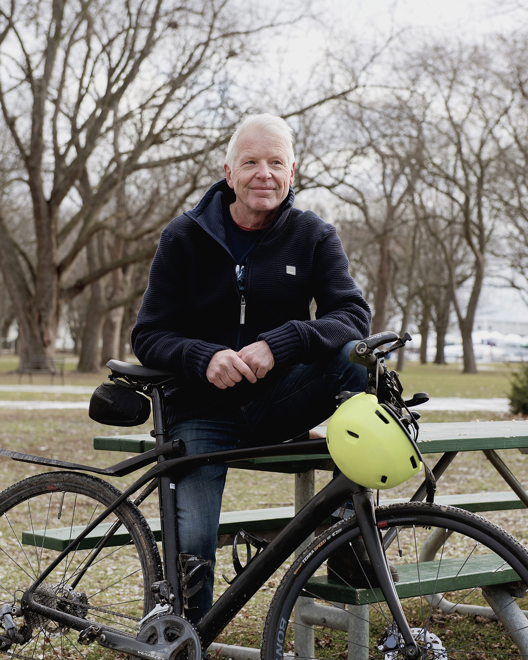 Professor Doug Richards is sitting on the edge a picnic table in a grassy field with trees bare of leaves in the background. His elbows are resting on the seat and handles of a mountain bike.