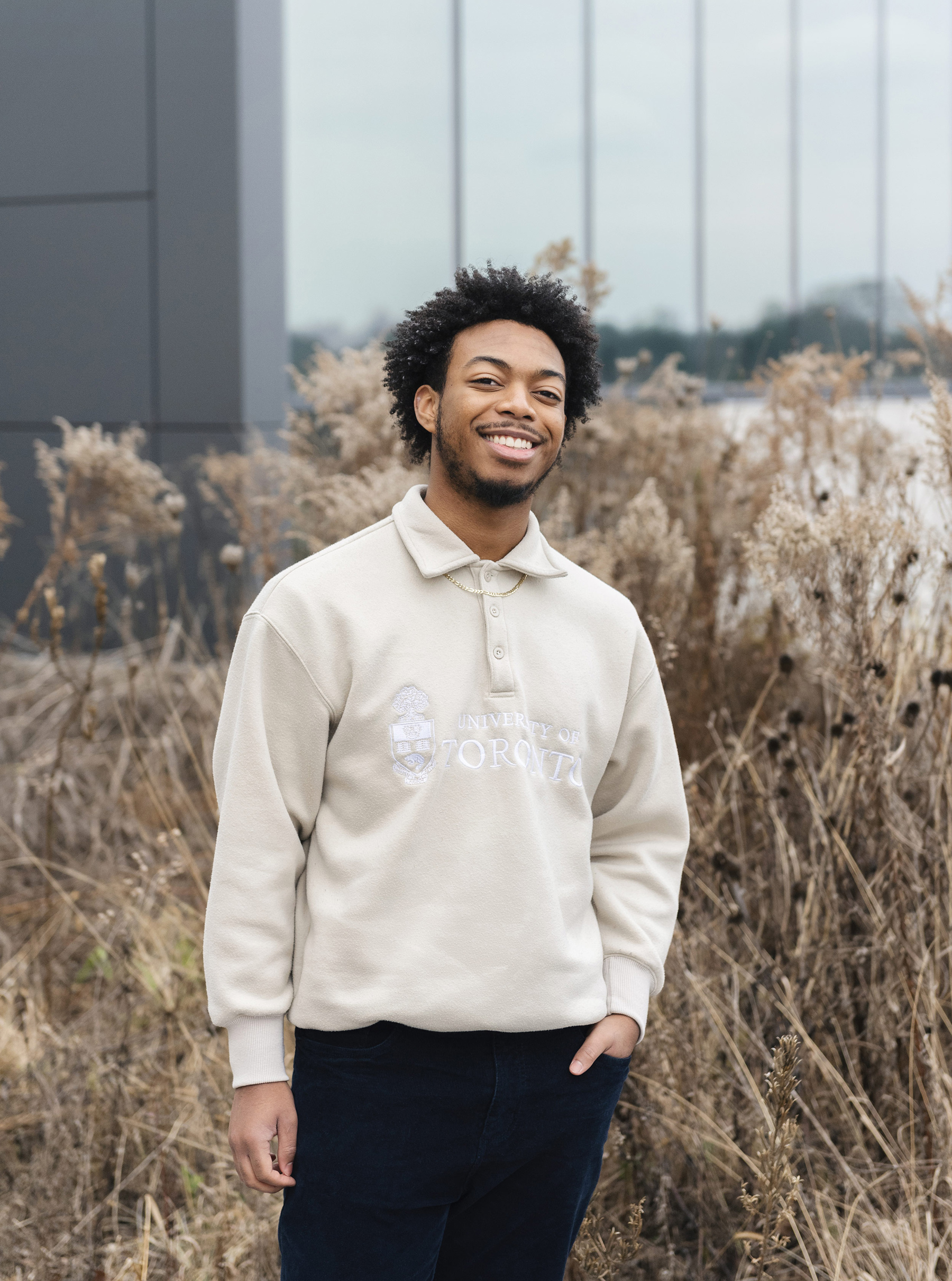 Outdoor shot of Giovanni Williams, wearing black jeans and a cream-coloured pullover with the University of Toronto logo embroidered on the front. He is standing in front of tall reedy shrubs, behind which is a building. The photo shows a partial view of the corner of the building, consisting of grey wall on the left and tall panelled windows on the right.