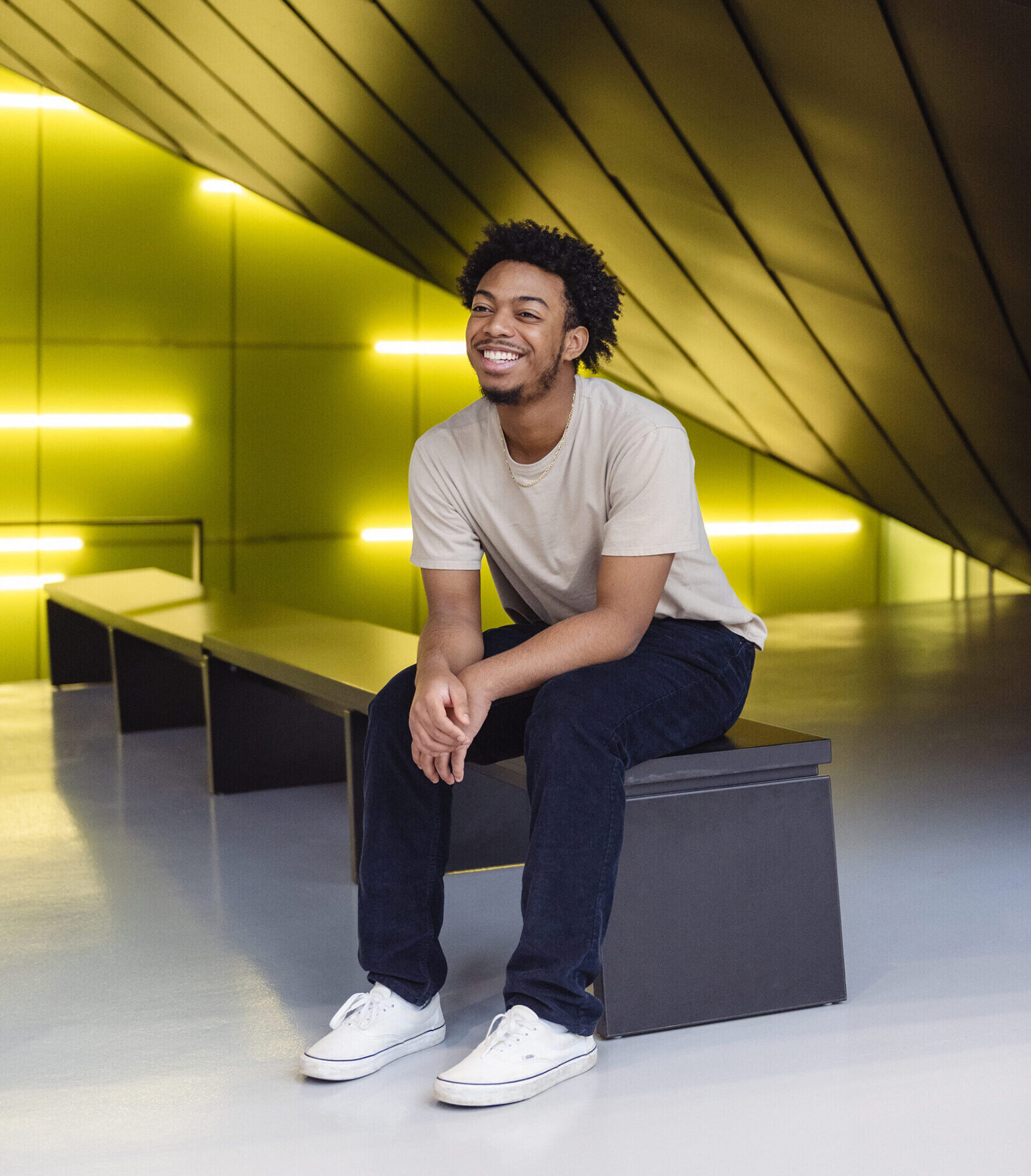 Giovanni Williams in a beige T-shirt, black jeans and white sneakers, sitting on a black wooden bench under a panelled ceiling that slopes down to the floor at the middle right of the photo. He is smiling and facing off camera, with his shoulders hunched and his forearms resting on his thighs. Several fluorescent tubes are placed along the wall at the far end of the bench, giving off a neon yellow light.