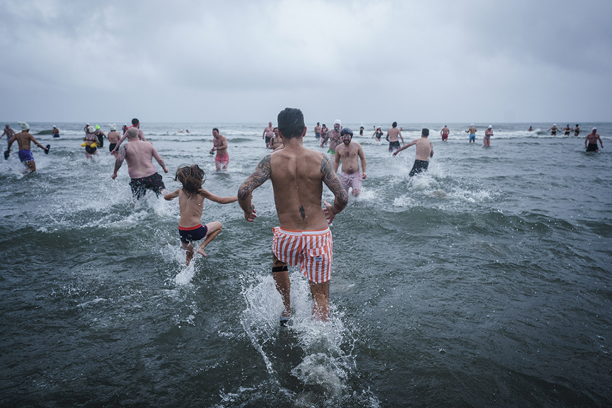 In the foreground, a man with tattooed arms and red and white striped swim trunks and a boy with long hair are splashing water as they walk into the surf. In the middle ground and background, people are standing or walking in the water. In the distance are the white foam of incoming waves.