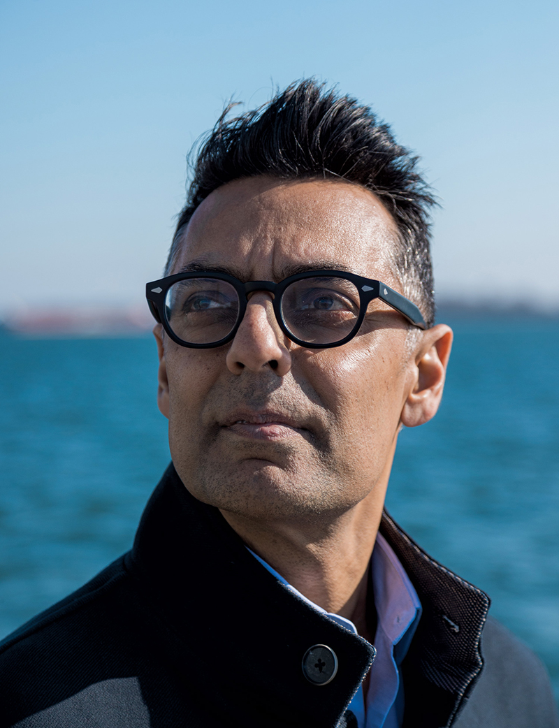 Headshot of Kamran Khan, wearing thick, black framed glasses and a dark jacket, looking off camera, with a body of water in the background