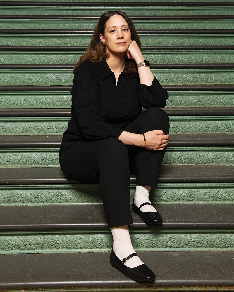Karina Bold, wearing a black, button-up blouse, black pants and black Mary Jane flats, sitting on a set of stairs with green, leaf-patterned moulding between dark grey steps