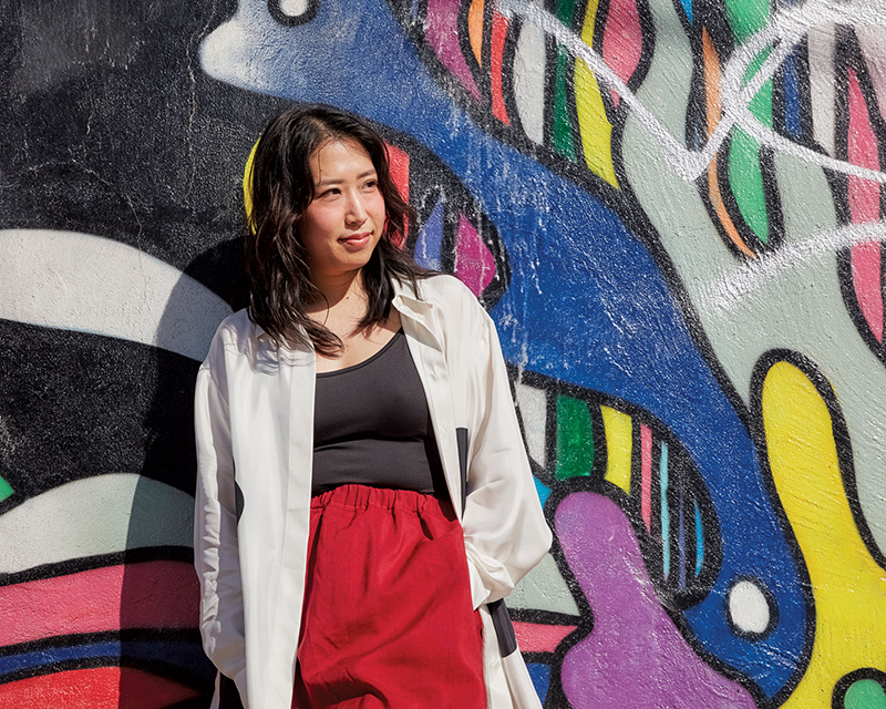 Professor Ai Taniguchi, wearing an open white button-up shirt over a black tank top and baggy red pants. She is outdoors, leaning against a graffiti covered wall.