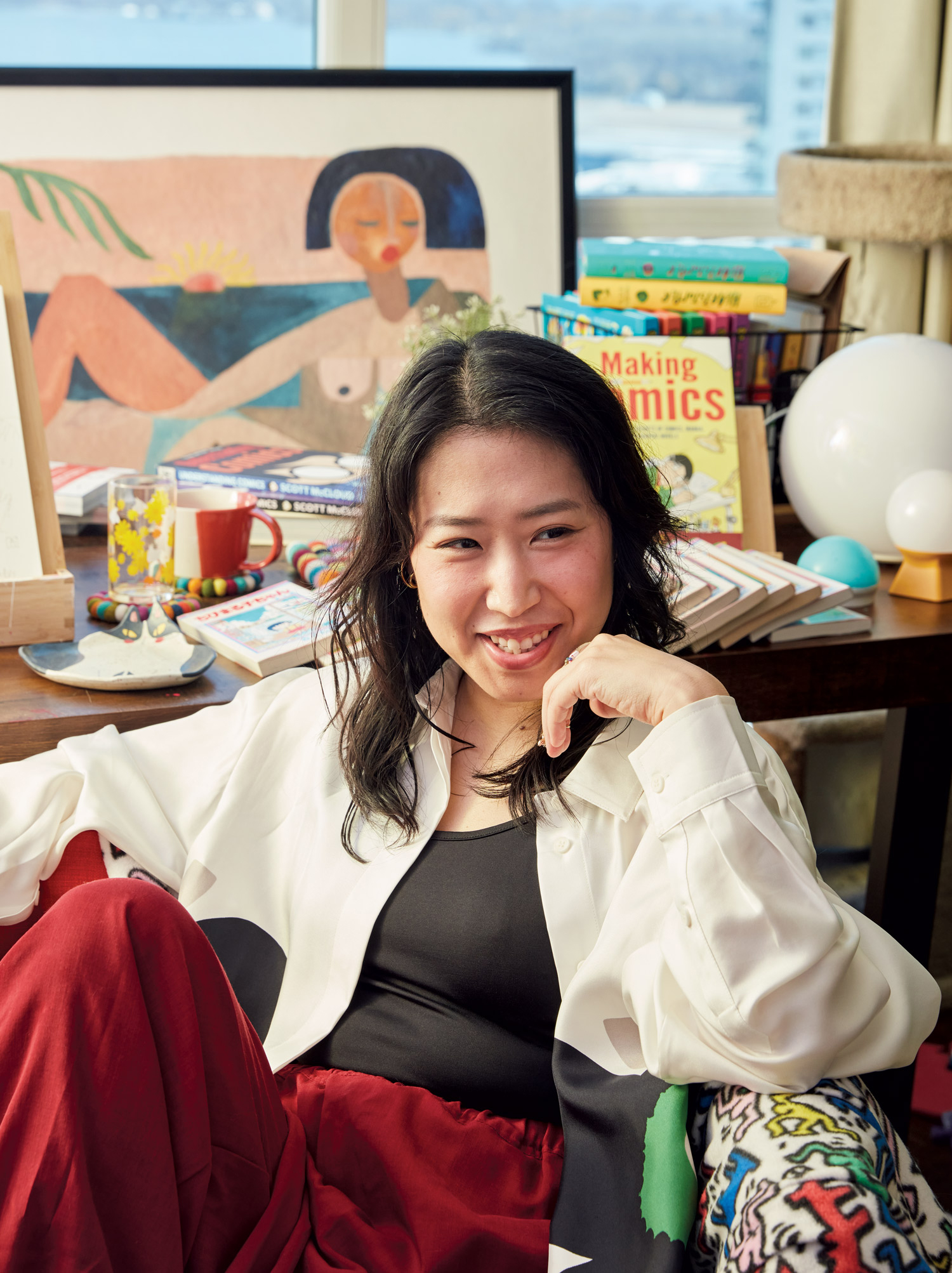 Professor Ai Taniguchi, wearing an open white button-up shirt over a black tank top and baggy red pants. She is sitting in an armchair, with an elbow against one arm of the chair, which is lined with a Keith Haring throw blanket. Behind her is a table full of books.