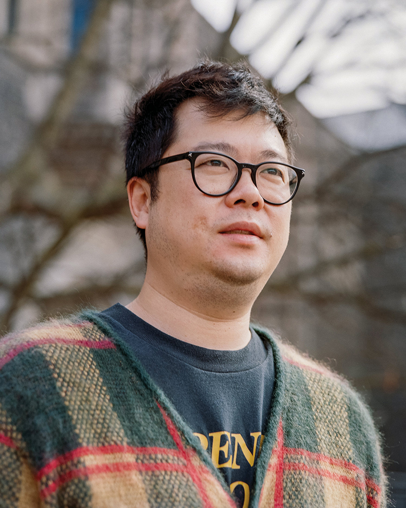 Outdoor headshot of Alex Wong, wearing black framed glasses and a green, yellow and red plaid cardigan over a sweatshirt. Behind him are bare tree branches out of focus.