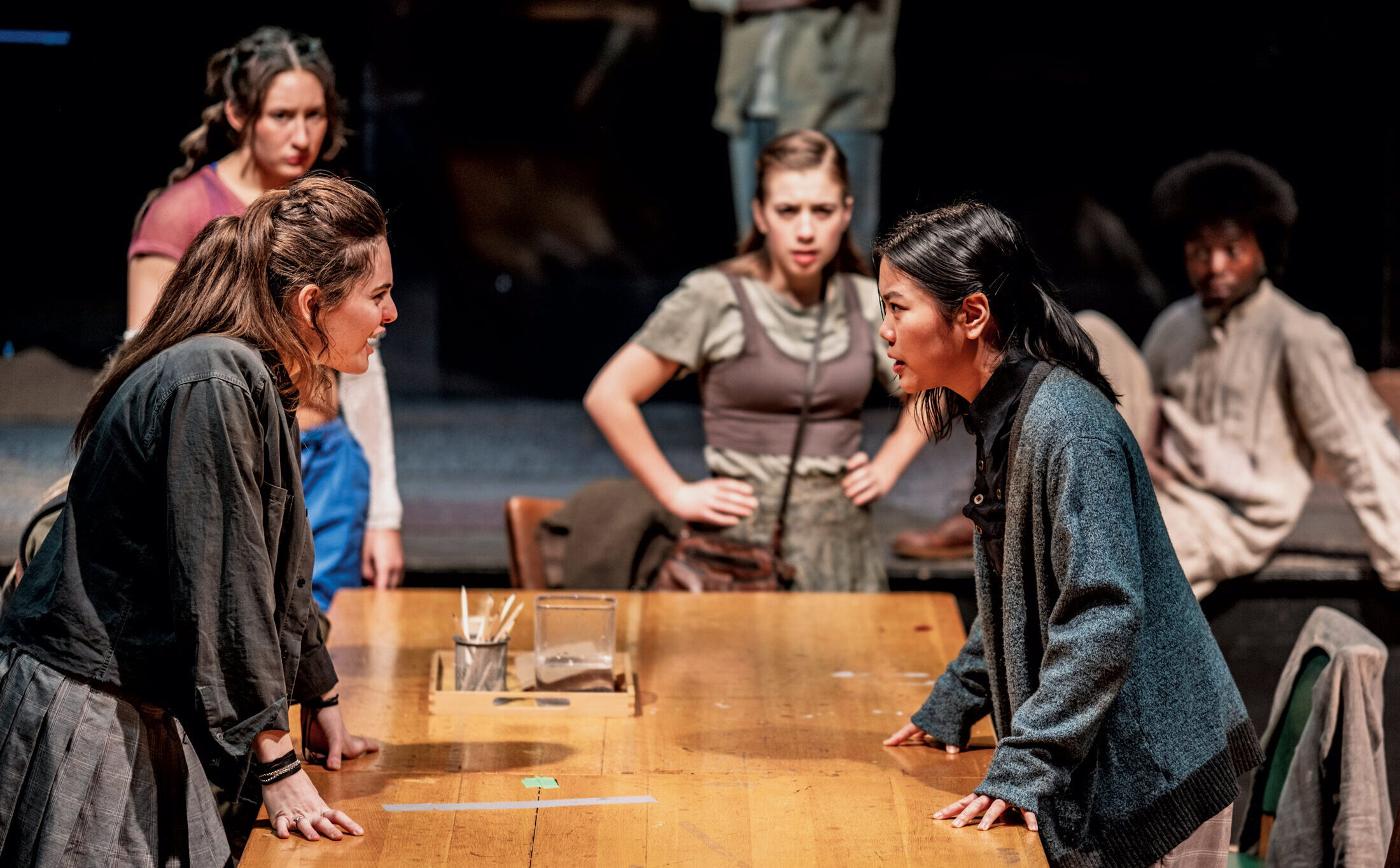 Students acting in a scene from The Trials. Two students appear to be in confrontation across a wooden table, with other students in the background, watching the pair.