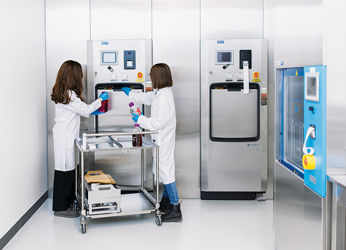 Evelina Tronina and Seema Malmji, wearing white lab coats and blue disposable gloves, are holding bottles of liquid, with a trolley between them. One student is pulling open a panel on the front of a tall, stainless-steel scientific device in a white lab room.