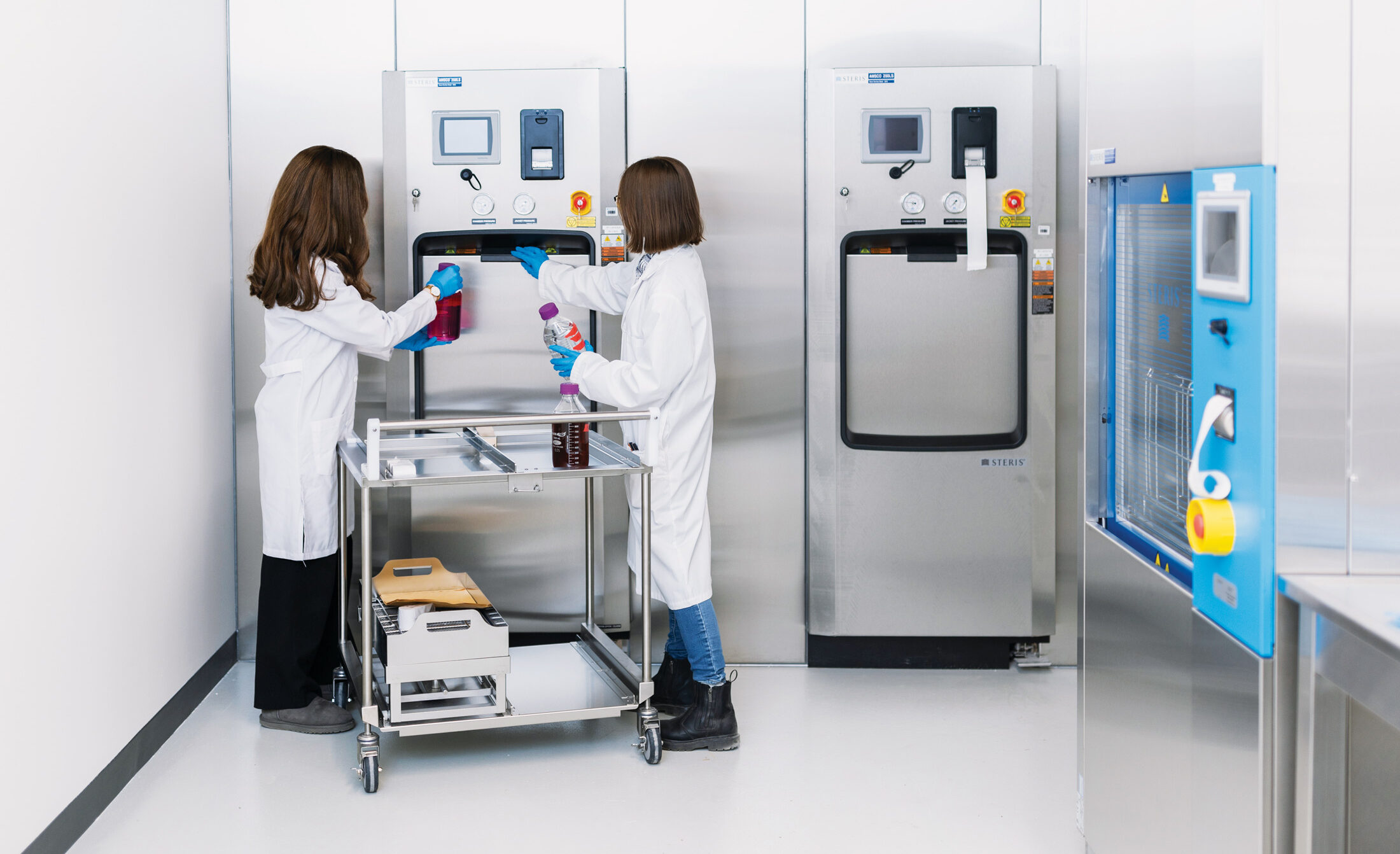 Evelina Tronina and Seema Malmji, wearing white lab coats and blue disposable gloves, are holding bottles of liquid, with a trolley between them. One student is pulling open a panel on the front of a tall, stainless-steel scientific device in a wet lab room.