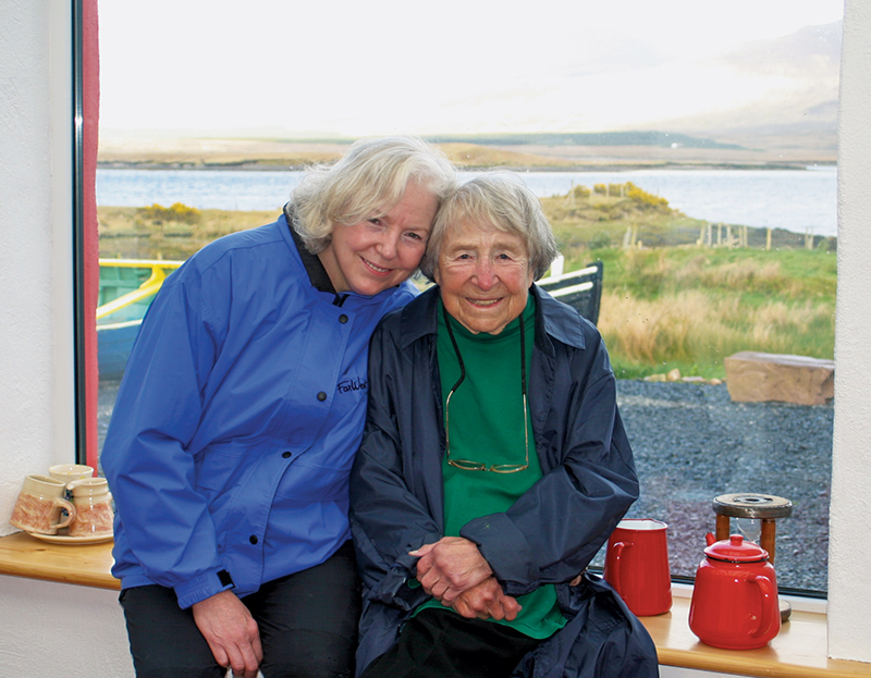 Wendy Wacko, in a blue windbreaker, and Doris McCarthy, in a navy blue windbreaker, green turtleneck and a pair of glasses hung around her neck, are sitting together on a wooden window sill by a large window overlooking pebbled ground, long grass and a body of water.
