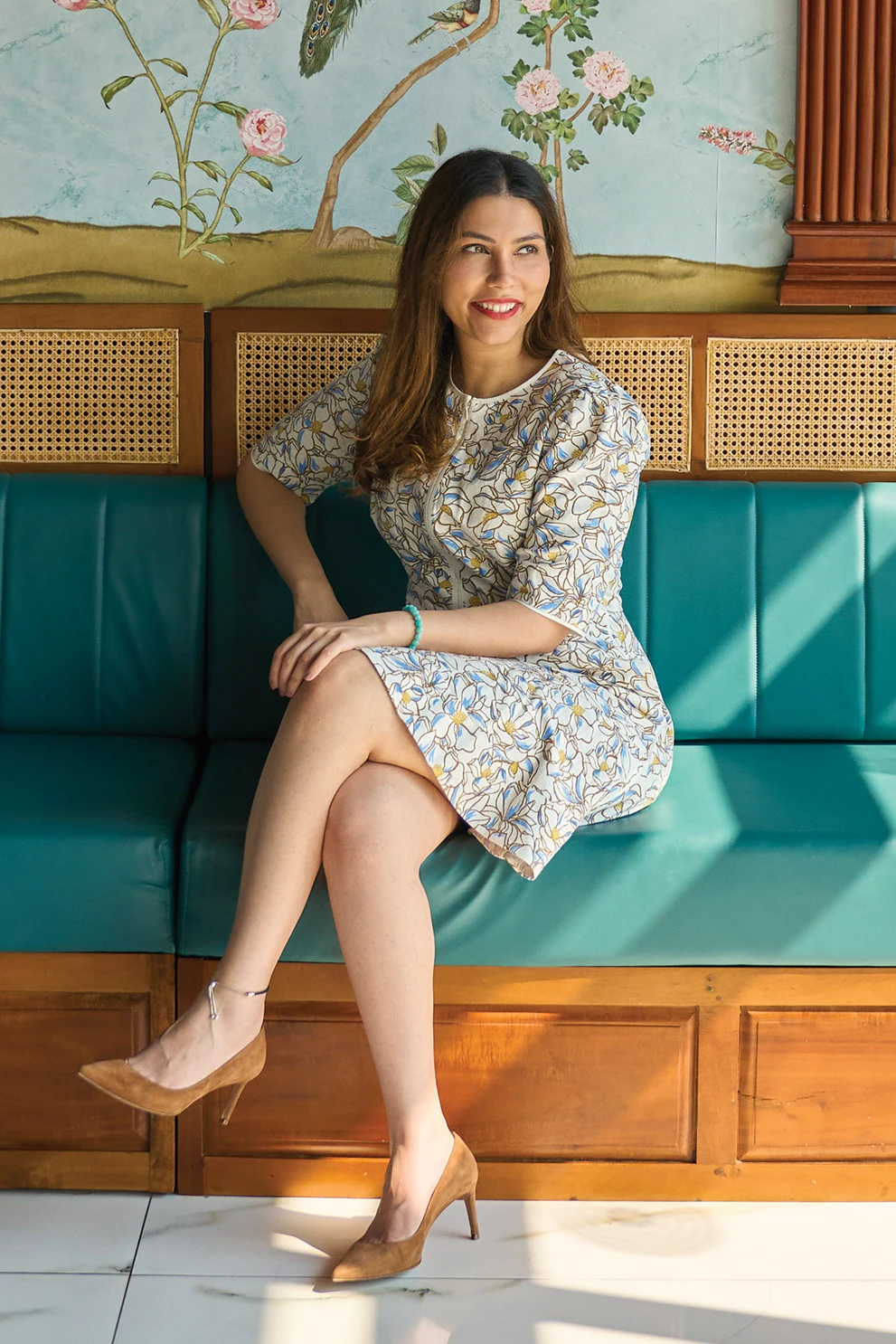 Ambica Jain in a flower-patterned dress, sitting on a green bench below a wall with a mural of pink flowers