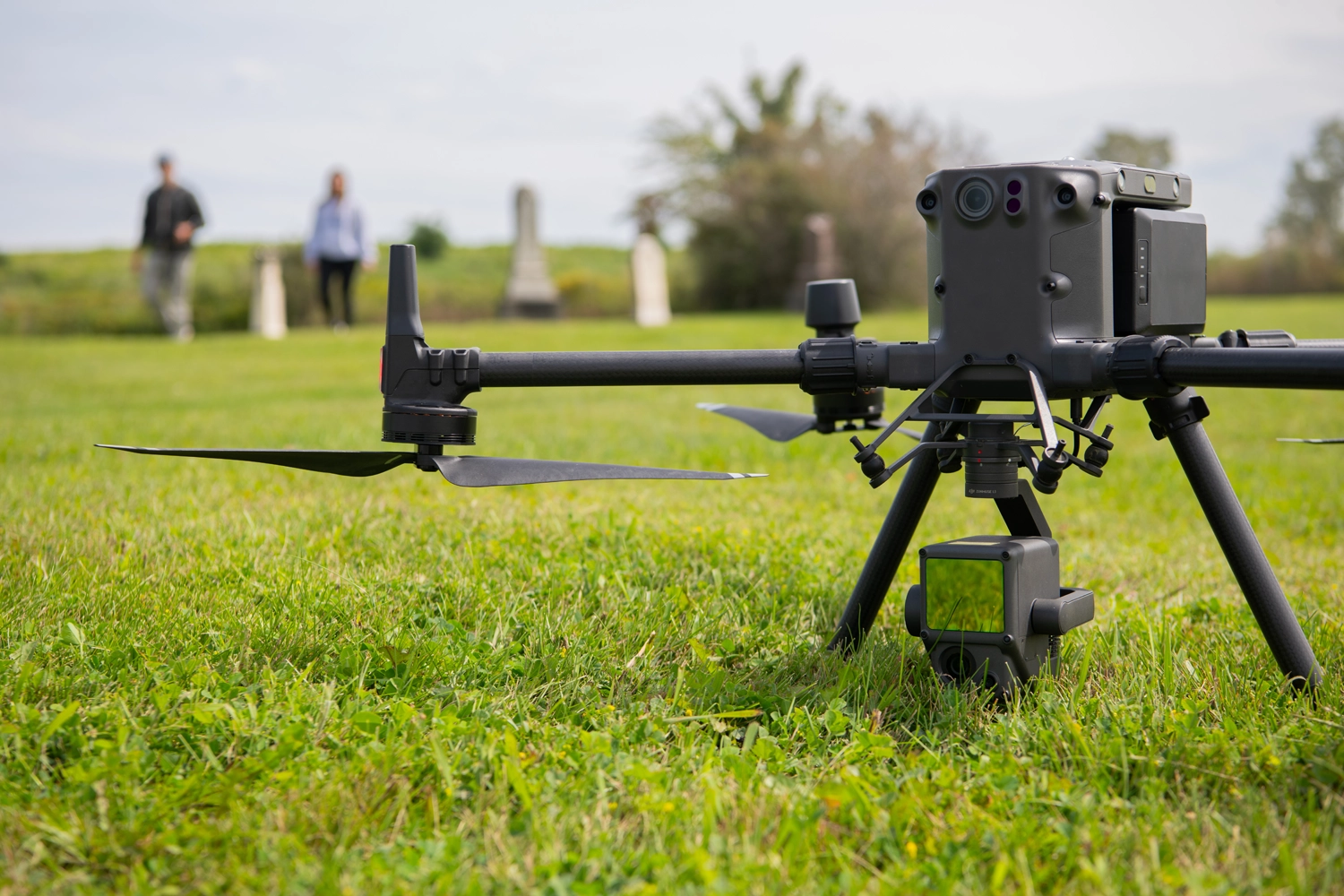 A close-up of a large drone sitting on a grassy field, with people and tombstones in the distance