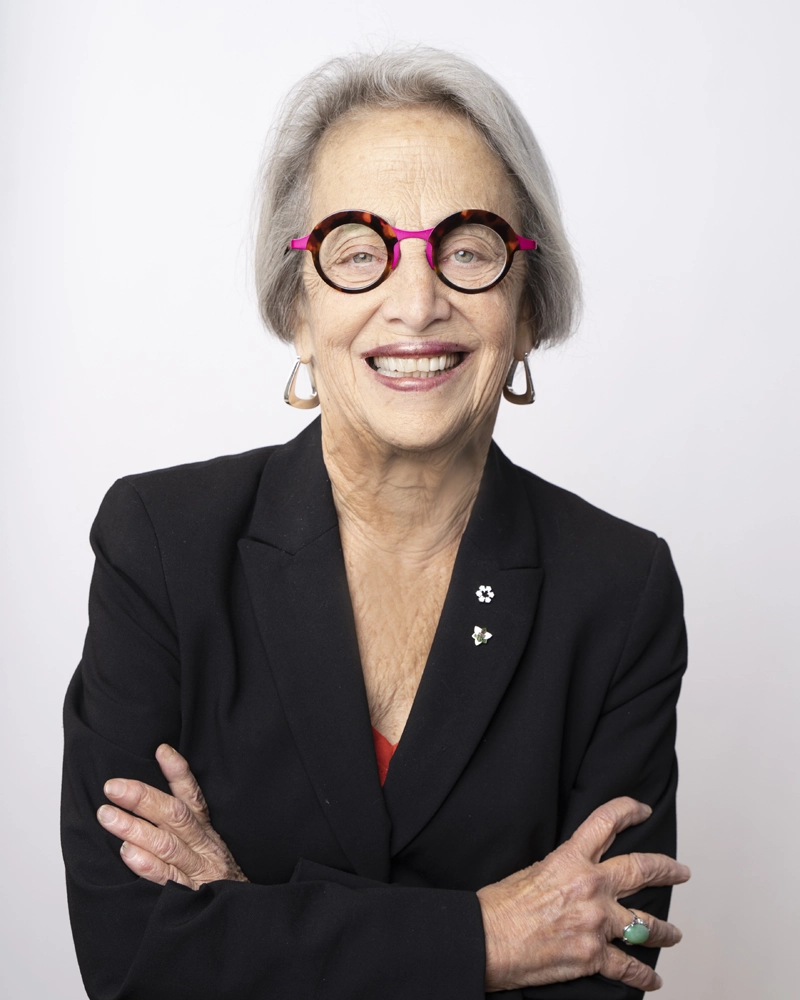 Professor Janice Stein, wearing thick magenta, orange and dark red framed glasses, standing with her arms crossed