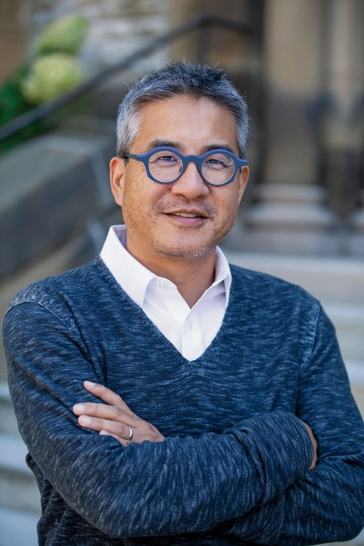 Outdoor photo of Professor Joe Wong in blue framed glasses and V-neck sweater, with his arms crossed