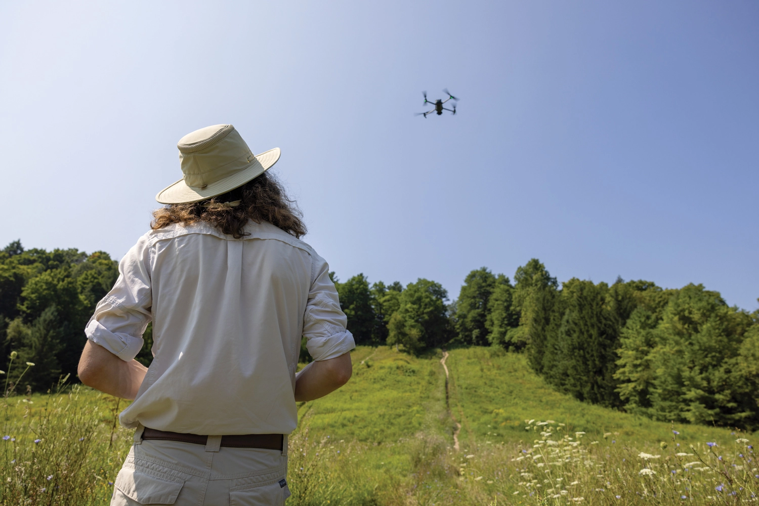 John English in a wide-brimmed hat, operating an aerial drone over an open field bordered by trees