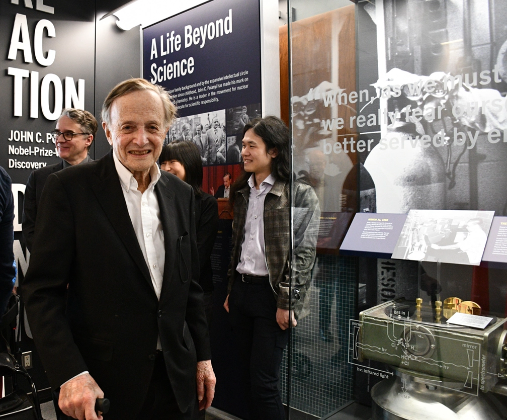 John Polanyi smiling and standing beside a glass display at an exhibit of his work