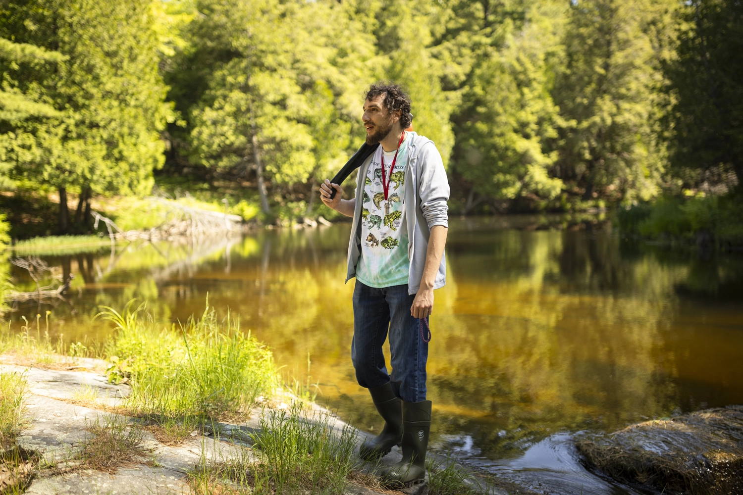 Allen Toulouse, wearing rubber boots, stands on the rocky shore of a still river