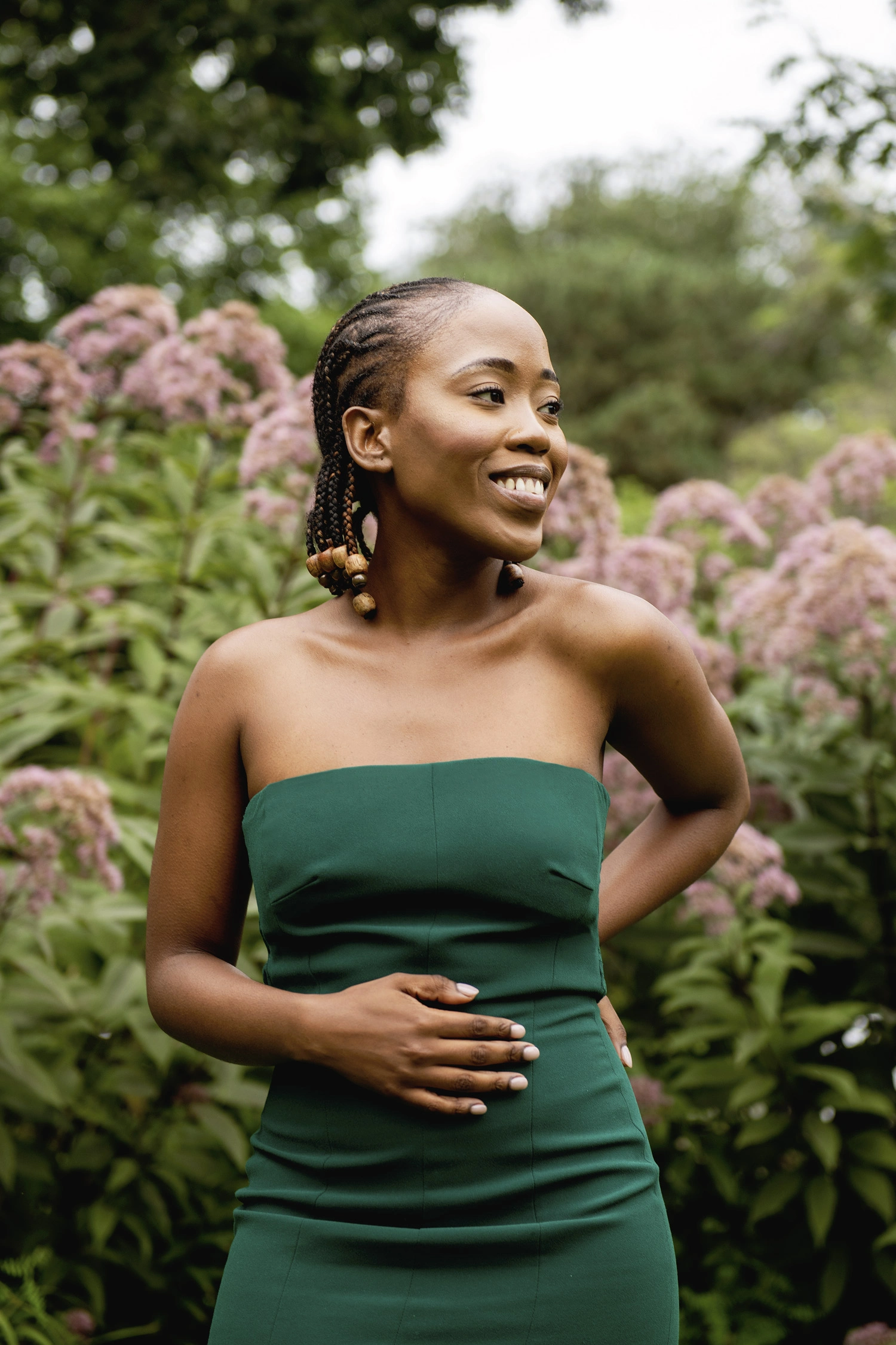 Outdoor photo of Mahlodi Letsie, wearing a green strapless gown and wooden beads at the end of her dreadlocks, standing in front of a flower bush