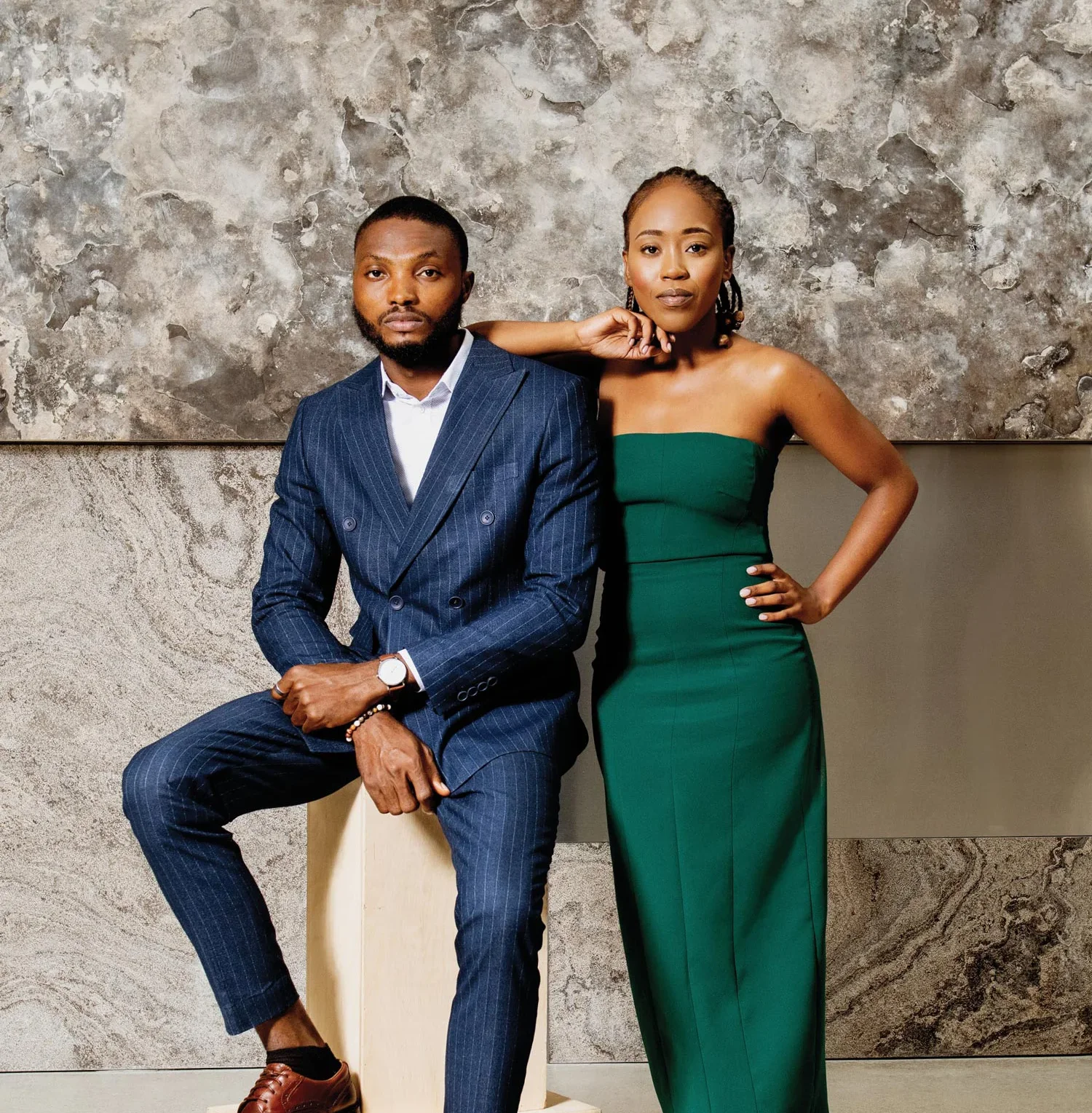 Mathew Okwoli in a blue, pin-striped suit, sitting on a wooden block on the left. Mahlodi Letsie in a green strapless gown standing on the right.