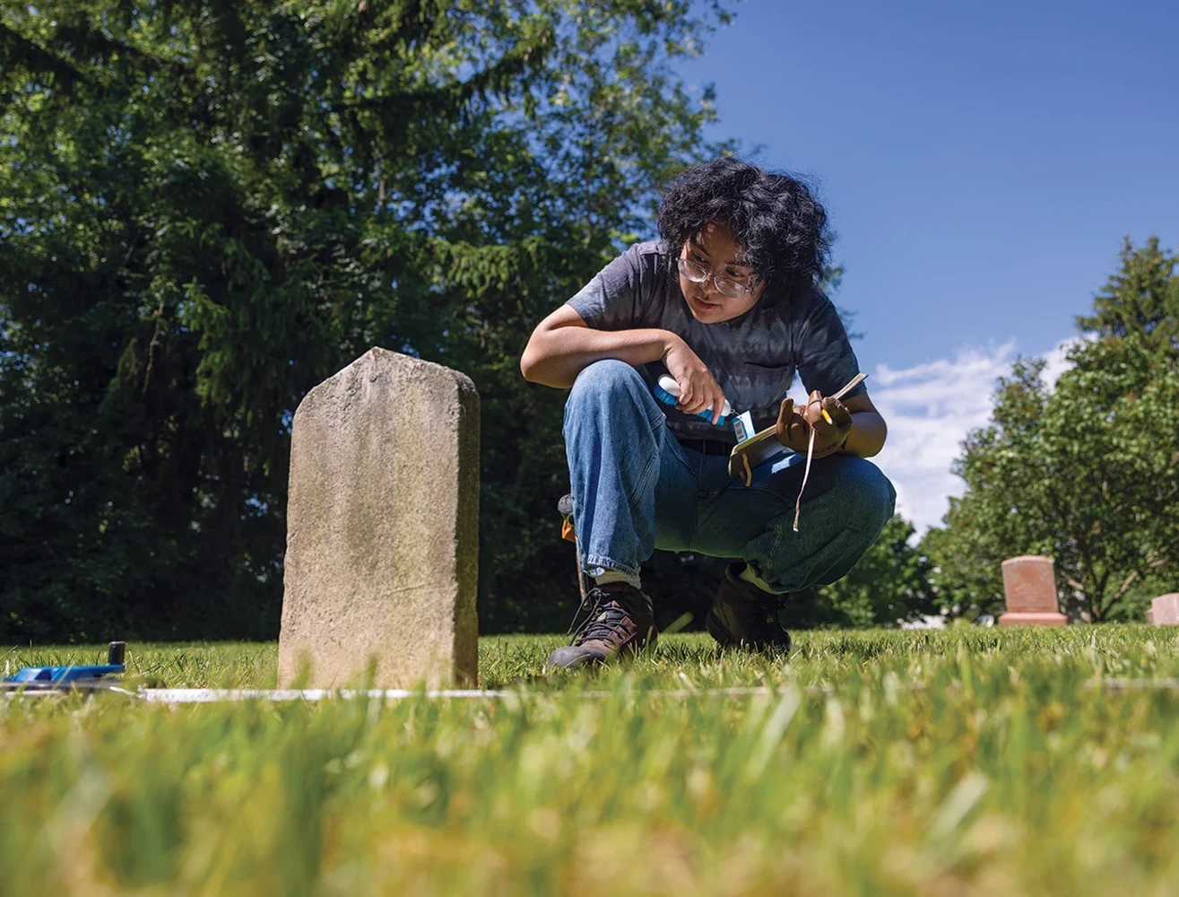 A researcher crouching down to examine a tombstone, an open notebook and pencil in one hand