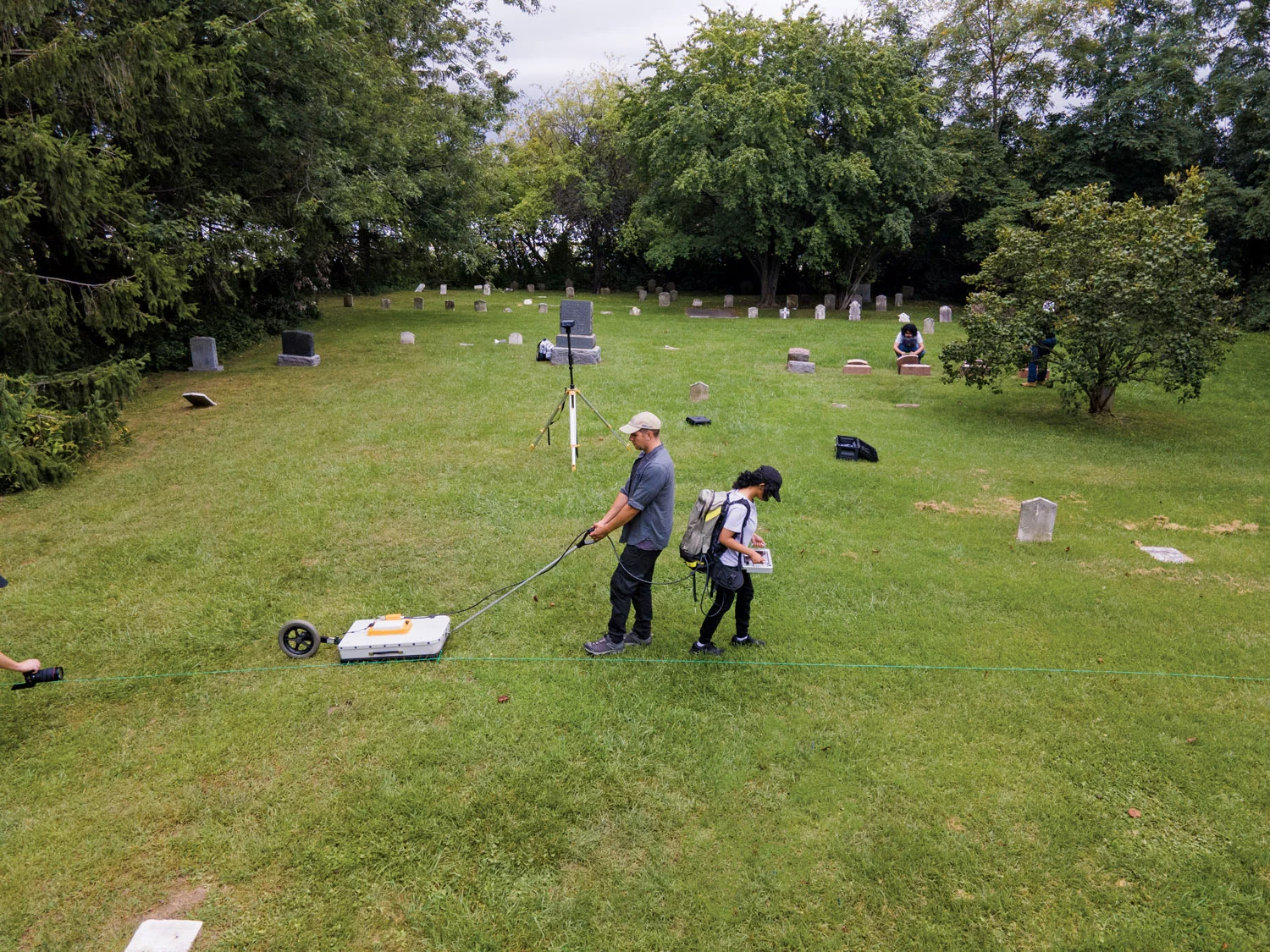 A researcher is pulling a ground-penetrating radar across a cemetery along a marked line. Another researcher is carrying a backpack and operating a monitor.