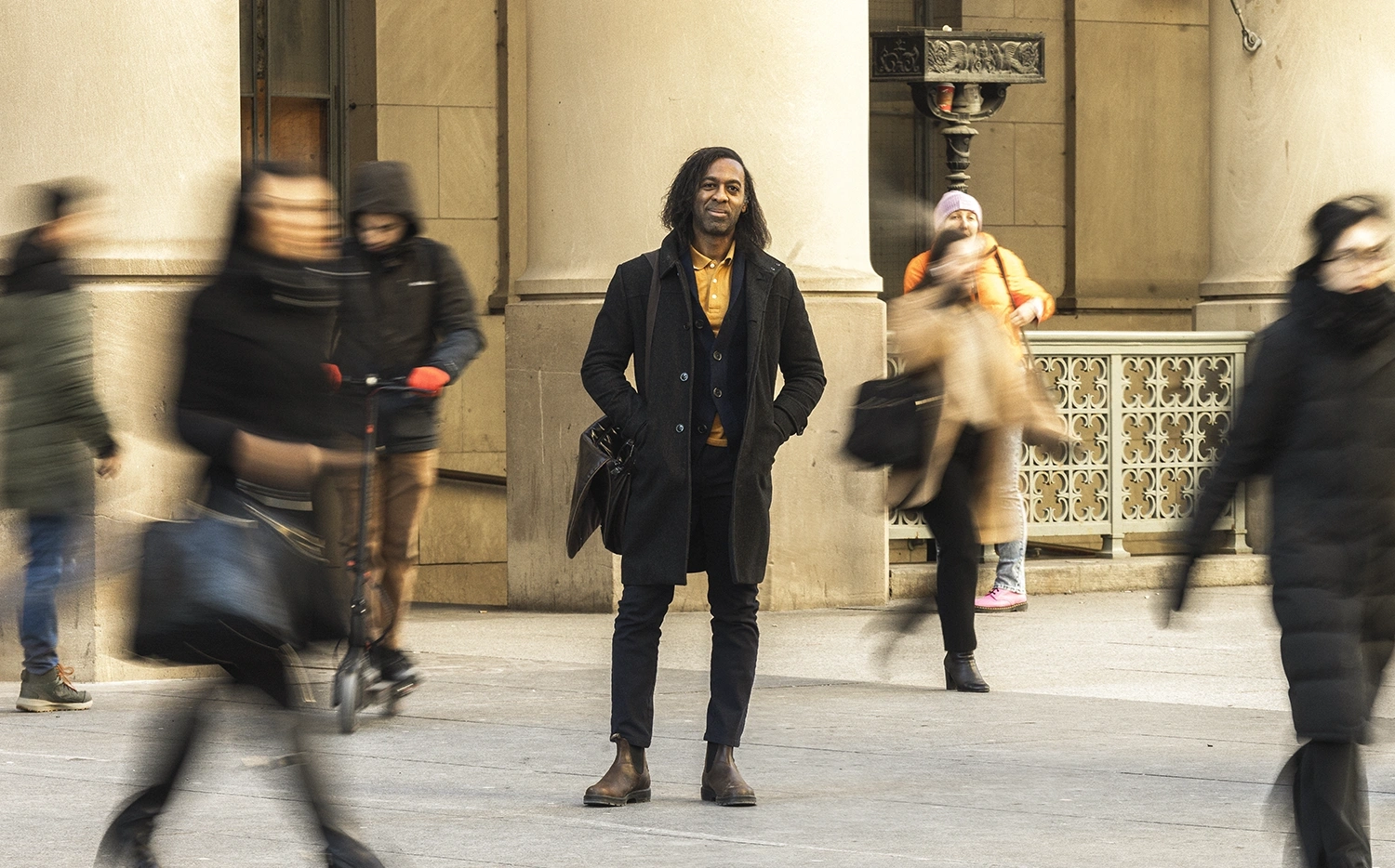 Professor Ian Williams standing still outside of a building with large columns, while people walking around him are motion blurred