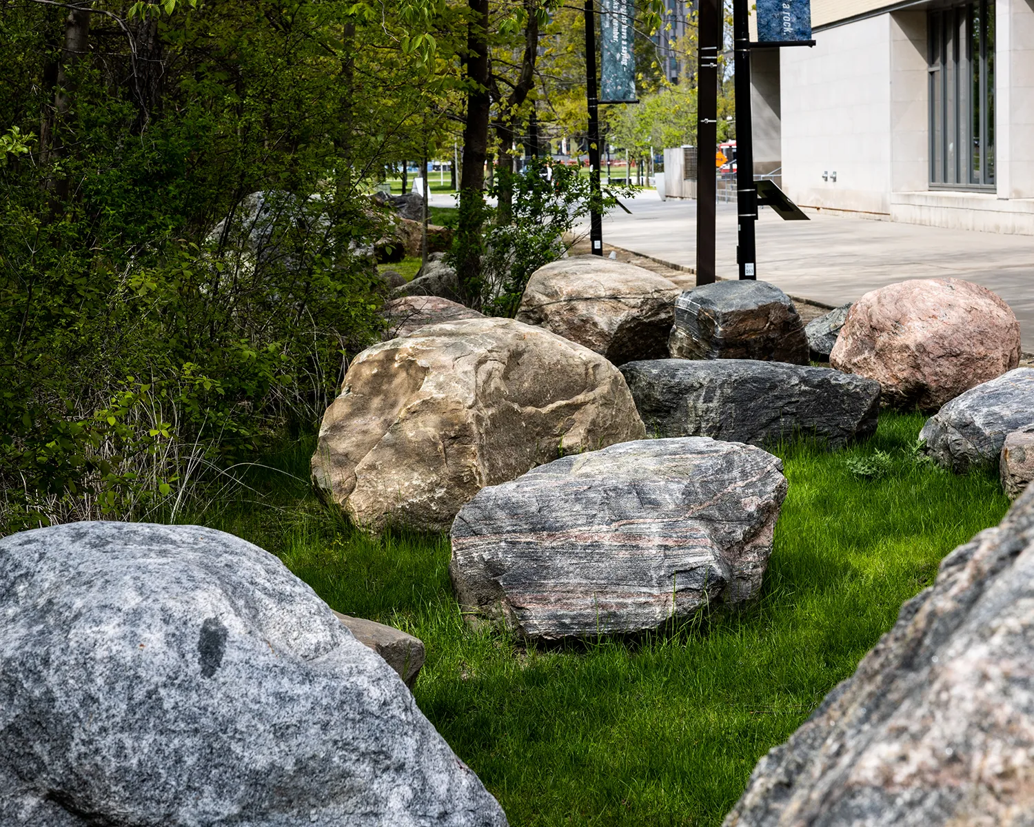 Different coloured granite boulders on grass, next to shrubs and a concrete pathway