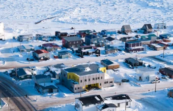 Bird's eye view of Inuusirvik Hub, a large light grey building with cutaways painted in yellow, among other smaller buildings, bordered by a field of snow