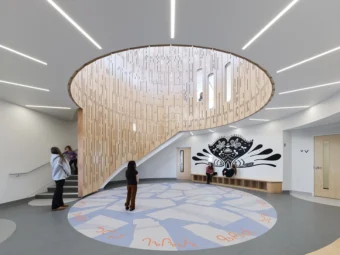 A few people in a circular white room with an Indigenous art mural on the far wall and a drum space with wood panelled siding partially extending down from the ceiling, bordering a set of stairs.
