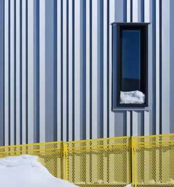 External, close-up view of a tall, narrow, black-framed window against the grey panelled side of the Inuusirvik Hub building, above a yellow fenced ramp