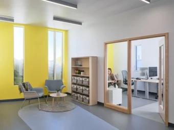 Angled view of an office. On the left is a sitting area next to a yellow wall with snow drifts visible through tall, narrow windows. On the right is a glass door leading to an enclosed space with desks and computers.