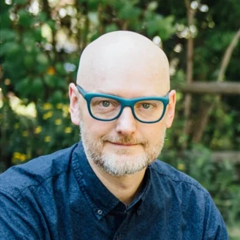 Headshot of Neil Macpherson, wearing thick blue-framed glasses and a blue denim shirt
