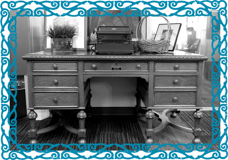 Front view of an antique wooden desk with a long drawer in the centre and three smaller drawers on each side. An old typewriter and other objects are placed on the surface.