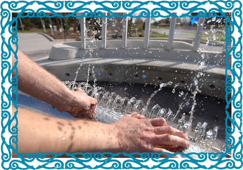 A pair of hands are placed with fingers touching a metal bar. Streams of water flow out from holes placed at regular intervals along the bar.