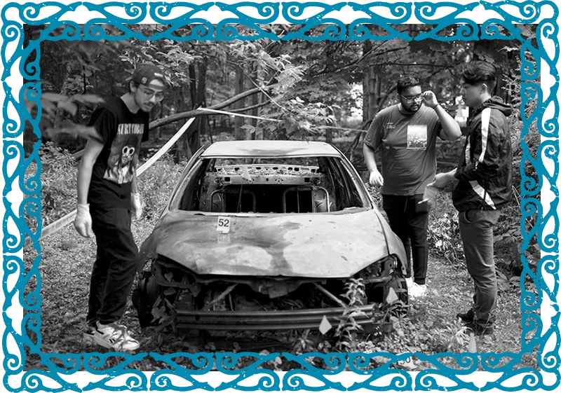 Three students wearing blue gloves examine a broken, rusted car in a forested area
