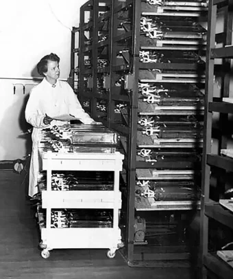 Black and white photo of Leone Farrell in a lab coat looking at items on a storage shelf, pushing a cart lined with glass bottles