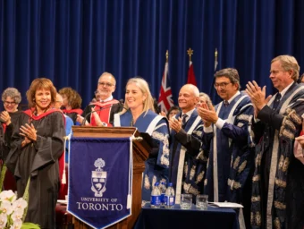 U of T President Melanie Woodin speaking onstage at a podium, standing beside past U of T presidents