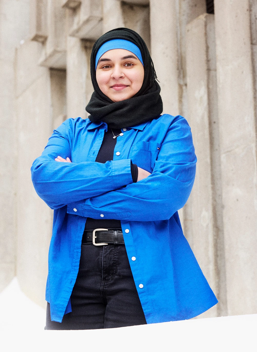 Dua Khan, wearing a bright blue unbuttoned shirt over a black top and a black head scarf over a bright blue hijab, standing with her arms crossed