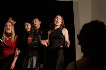 A diverse group of five students singing against a black backdrop, the shadowed head of an audience member in the foreground