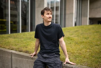 Velislav Dzhebarov, in a black T-shirt and grey pants, sitting on a concrete border with a field of grass behind him, smiling and looking off to the side